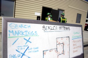 A search and rescue plan is explained on a white board as CERT members wait their turn to search a dark building while crawling on their hands and knees and working as a team.