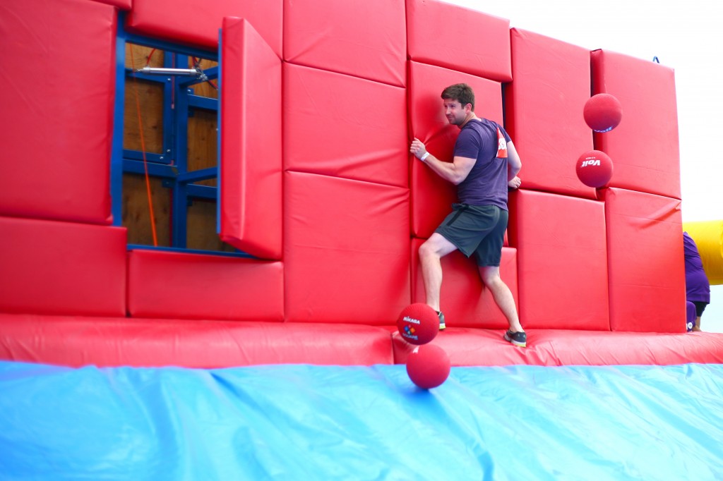 Carefully making his way across the Whacking Wall, a man holds on to one of the sections that pop out as he gets bombarded with red bouncy balls.