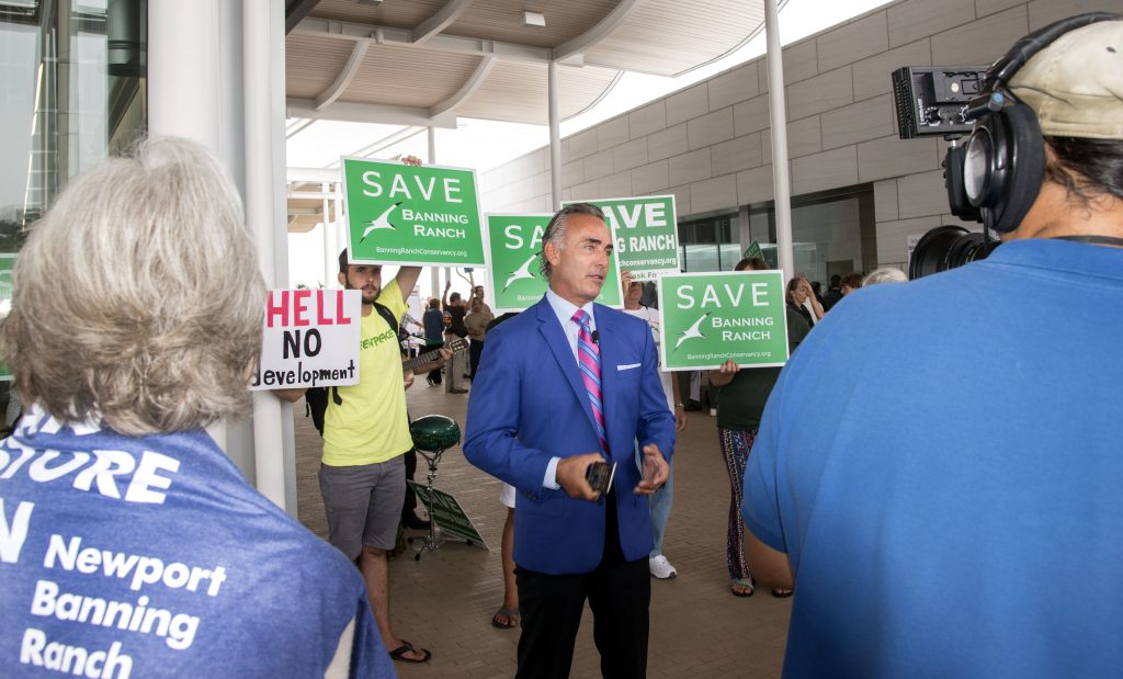 A Newport Banning Ranch project supporter talks to the press Wednesday as opponents wave signs behind him. — Photo by Charles Weinberg ©