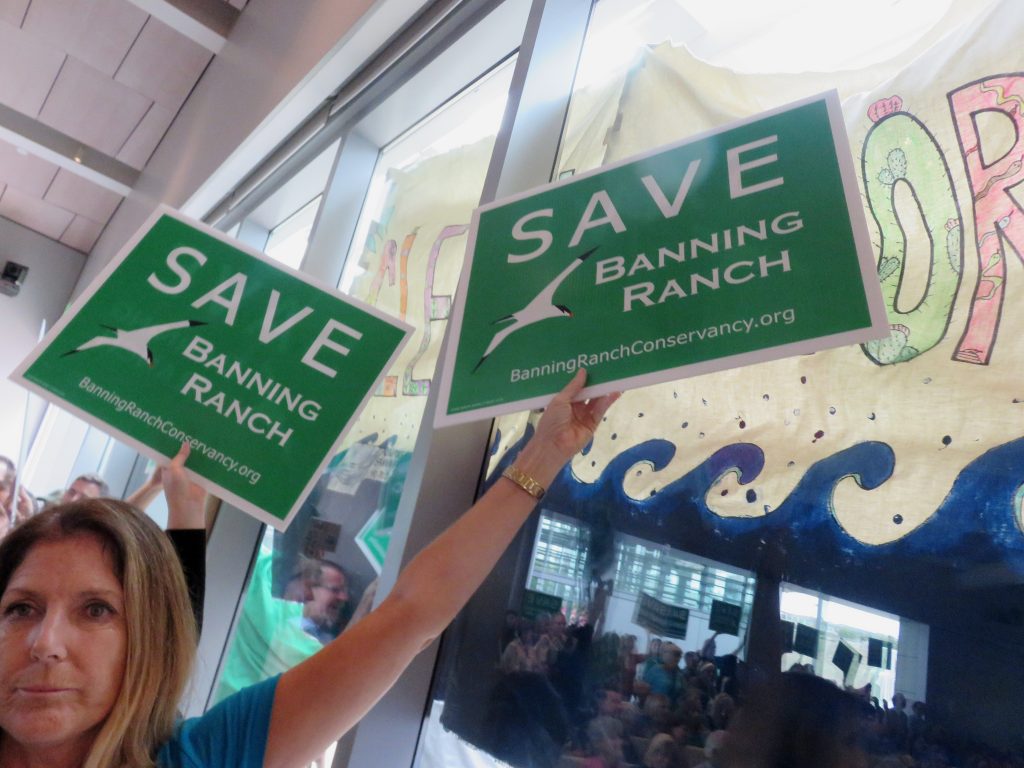 An opponent to the project holds a "Save Banning Ranch" sign in front of an NBR supporter's "Clean. Restore. Open." banner, while the audience is reflected in the glass, during the California Coastal Commission meeting Wednesday. — Photo by Sara Hall ©