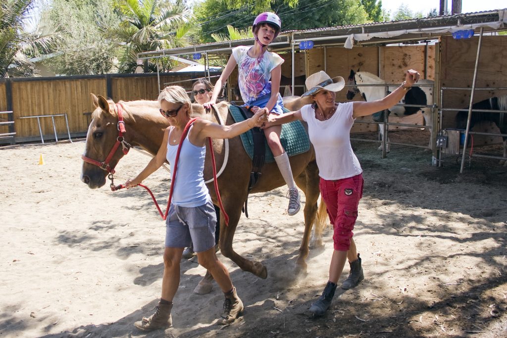 Michelle Hoops looks over at the other horses as she rides Peanut, a 19-year-old quarter horse. Bernadette Olsen (right, in cowboy hat), Vickie Tombrello (front left) and Katharina Chiu lead the horse and rider around the arena at the Back Bay Therapeutic Riding Club during Hoops’ July 14 riding session. — Photo by Sara Hall ©