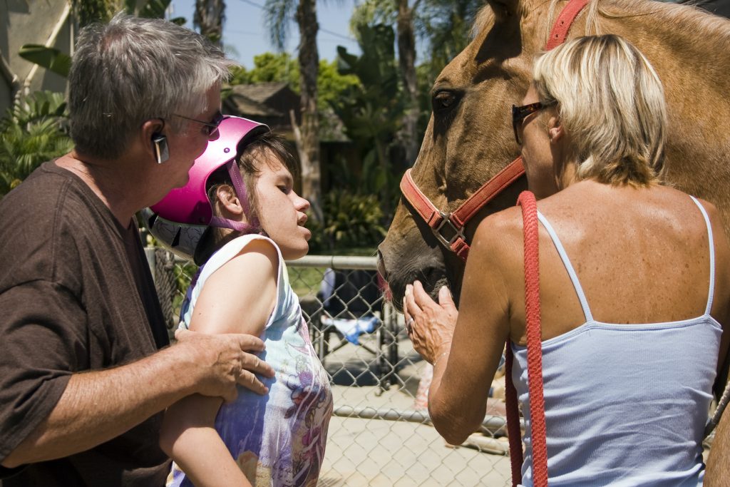 Michelle Hoops gets close to her favorite horse, Peanut, with the assistance of her dad, Mike, at Back Bay Therapeutic Riding Club (BBTRC) in Newport Beach before hand-feeding him a carrot. Horse manager Vickie Tombrello stands with Peanut as he gently takes the treat from Michelle’s hand. — Photo by Sara Hall ©